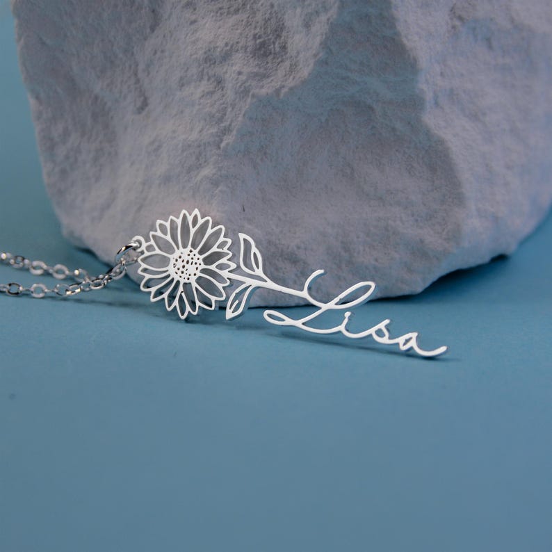 May include: A silver sunflower pendant necklace with the name "Lisa" in a cursive font. The necklace features a delicate sunflower design and is displayed against a blue background and a textured gray stone.