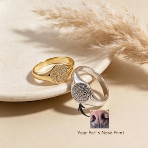 Puede incluir: Dos anillos tipo sello, uno dorado y otro plateado, cada uno con una cara circular de superficie texturizada. Una flecha señala desde el anillo plateado hacia una pequeña imagen insertada de una nariz de perro con el texto Your Pet's Nose Print.