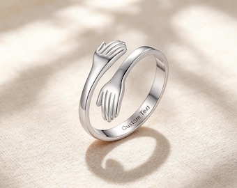 May include: A silver-toned open ring featuring two hands that meet in the centre to form a hug design. The band of the ring includes the engraved text Custom Text on the outer surface, showcasing a polished metallic finish throughout.