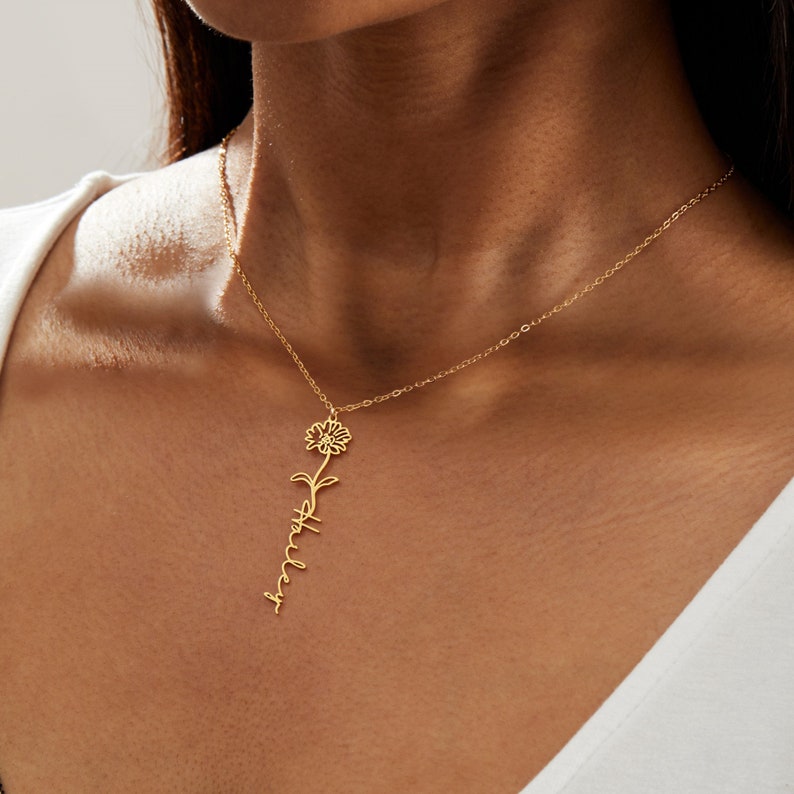 May include: A gold-colored necklace featuring a floral pendant with the name "Ashley" written in a delicate script. The necklace has a thin chain and is displayed against a neutral background.
