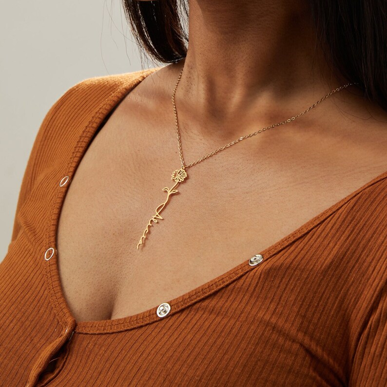 May include: A gold-colored necklace featuring a sunflower design with the name "Anna" written in a delicate script. The necklace is suspended from a fine chain. The person in the image is wearing a brown long-sleeved shirt.