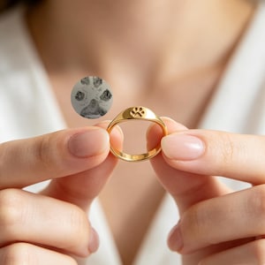 以下が含まれることがあります： 平らな円形の面に小さな肉球の刻印が施されたゴールドトーンのシグネットリング。リングの上には、明るい背景にダークグレーのインクスタイルで描かれた詳細な動物の肉球のインプレッションを表示する円形のグラフィックが浮かんでいます。