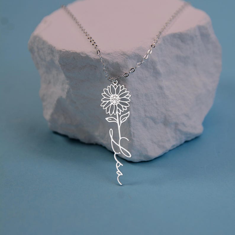 May include: A silver-colored necklace featuring a sunflower pendant with the name "Lisa" written in cursive script below the flower. The necklace is displayed on a white, textured surface against a blue background.