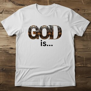May include: White t-shirt featuring the word "GOD" in a textured font, with a church image within the letters. Beneath, the words "is..." are printed in black. The shirt is shown on a wooden surface.
