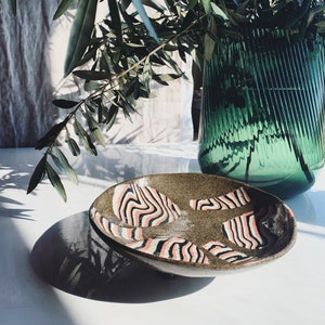 Pode incluir: Uma tigela de cerâmica artesanal com base castanha salpicada e padrões de ondas abstratas rosa, pretas e brancas. A tigela oval está sobre uma superfície branca, com um vaso de vidro verde e ramos de oliveira ao fundo.