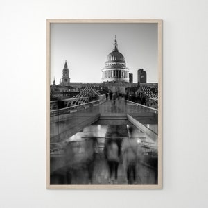 Può includere: Una fotografia in bianco e nero della Cattedrale di San Paolo a Londra, in Inghilterra. La cattedrale è vista da un ponte, con persone che camminano sul ponte in primo piano. La foto è scattata da un angolo basso, guardando la cattedrale.