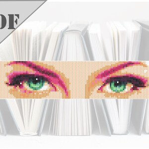 Puede incluir: Primer plano de un patrón de punto de cruz que representa un par de ojos con iris verdes y sombra de ojos rosa.