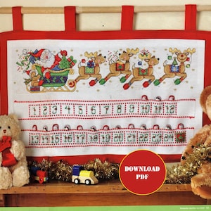 Puede incluir: Un calendario de adviento de tela blanca con un borde rojo y un diseño de punto de cruz de Santa Claus en un trineo tirado por renos. El calendario tiene 24 bolsillos numerados para golosinas.