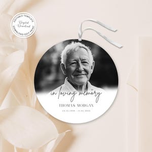 May include: A round, black and white memorial ornament featuring a portrait of an elderly man. The text "in loving memory" and the name "Thomas Morgan" are displayed, along with the dates 12.13.1950 - 11.01.2025. A white ribbon is attached for hanging.