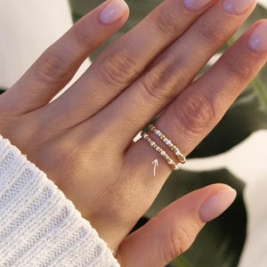 Könnte beinhalten: Zwei zarte goldene Ringe, jeder mit kleinen, abwechselnd weißen und goldenen Perlen verziert. Die Ringe sind auf einem Finger gestapelt und zeigen ein minimalistisches Schmuckdesign. Die Hand trägt einen weißen Pullover, im Hintergrund eine grüne Pflanze.