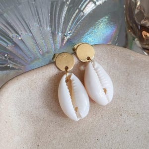 Può includere: Orecchini a bottone color oro con pendenti a conchiglia bianca. Gli orecchini hanno un bottone rotondo dorato e un piccolo anello dorato che si collega alla conchiglia. Le conchiglie sono bianche con una superficie naturale e strutturata.