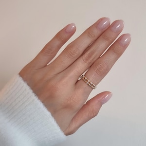 Könnte beinhalten: Ein zarter Ring bestehend aus zwei dünnen Bändern, die mit kleinen, schillernden, hellen Perlen und winzigen goldfarbenen Metallkugeln verziert sind. Der Ring wird am Ringfinger getragen und zeigt eine subtile, schimmernde Textur und ein raffiniertes Design.