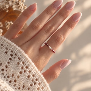 Könnte beinhalten: Ein zarter Ring bestehend aus einer Abfolge kleiner, dunkelroter Perlen, die mit winzigen metallisch silbernen Perlen und einer einzelnen weißen Perle in der Mitte durchsetzt sind. Der Ring wird am Finger getragen und zeigt den Kontrast der Farben.