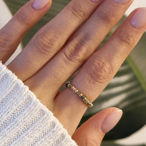 Könnte beinhalten: Ein goldener Perlenring mit grünen, braunen und klaren Perlen. Der Ring befindet sich an einem Finger, mit einem weißen Ärmel im Bild. Die Person hat hellrosa Nagellack.