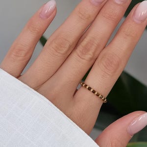 Könnte beinhalten: Ein dünner Ring mit einem sich wiederholenden Muster aus kleinen, abwechselnden goldfarbenen Metallperlen und dunkelbraunen runden Perlen. Das Band ist kreisförmig und zeigt ein filigranes Design mit einer gleichmäßigen Textur.