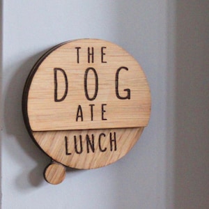 May include: A round wooden sign with the text "THE DOG ATE LUNCH" in black lettering. The sign is made of light brown wood and has a small wooden knob on the bottom.