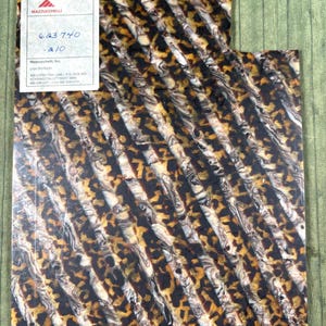 May include: A rectangular sheet of cellulose acetate material featuring a repeating pattern of diagonal stripes in shades of brown, black, and cream. A white label in the top left corner displays the text Mazzucchelli, 623 740, and 210.