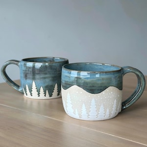 May include: Two ceramic mugs featuring a blue glazed upper section and a cream-colored lower section. The lower portion is decorated with a white forest of pine trees and small dots resembling falling snow, creating a winter landscape design.