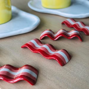 May include: A collection of ceramic bacon-shaped pieces, featuring red, pink, and white stripes, are displayed on a light brown surface. These glossy pieces are designed to look like bacon rashers. White plates and yellow objects are also visible.