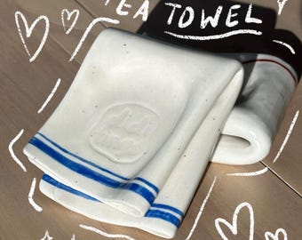 Może przedstawiać: Złożony, kremowy ceramiczny uchwyt na ściereczkę kuchenną o nakrapianej fakturze, z dwoma poziomymi niebieskimi paskami przy dolnej krawędzi i wytłoczonym logo na środku. Nad przedmiotem znajduje się biały napis "TEA TOWEL".