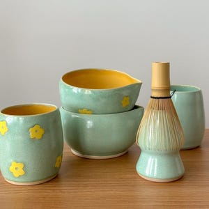 Może przedstawiać: Zestaw pięciu jasnozielonych ceramicznych misek z ż&oacute;łtymi akcentami kwiatowymi. Miski są ułożone w stos i ustawione na drewnianej powierzchni. W zestawie znajduje się bambusowa trzepaczka.