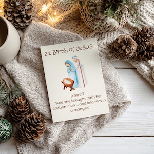 May include: A Christmas card with the text "24. Birth of Jesus" and a watercolor illustration of the Nativity scene. The card is surrounded by pine cones, greenery, and a neutral-colored blanket.