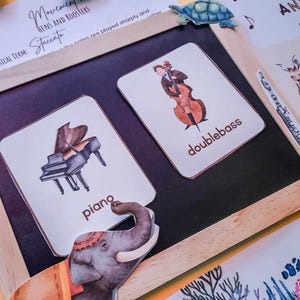 May include: A set of flashcards featuring illustrations of musical instruments, including a piano, double bass, and violin. The cards are on a chalkboard with a wooden frame. The text on the cards reads "piano", "double bass", and "violin".