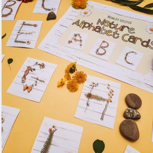 Op de afbeelding: Alfabetkaarten met letters gemaakt van natuurlijke elementen zoals bloemen en takjes. De kaarten zijn gerangschikt op een gele ondergrond, met extra bloemen en kleine stenen. De tekst "Nature Alphabet Cards" is zichtbaar.