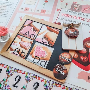 May include: A Valentine's Day themed educational set. A wooden frame displays alphabet letters A-D with heart-shaped cookies. Additional elements include word cards, a decorative ornament, and number cards. The background features pink and white stationery.