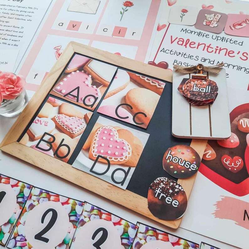 May include: A Valentine's Day themed educational set. A wooden frame displays alphabet letters A-D with heart-shaped cookies. Additional elements include word cards, a decorative ornament, and number cards. The background features pink and white stationery.