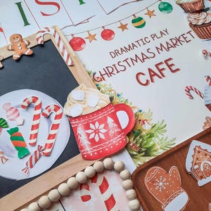 May include: A white sign with the text "Dramatic Play Christmas Market Cafe" in red and green lettering. The sign is surrounded by various Christmas-themed decorations, including candy canes, gingerbread cookies, and a red mug with marshmallows.