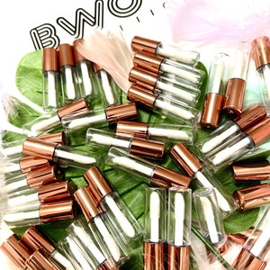 May include: A large group of clear glass tubes with rose gold lids and white applicators. The tubes are arranged in a pile on a green and white surface.