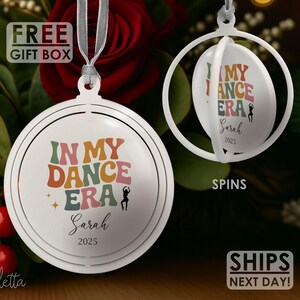 May include: White, round Christmas ornament with the text "IN MY DANCE ERA" in retro colors. The ornament includes a dancer silhouette, the name "Sarah", and the year "2025". A second ornament spins to reveal the same text. Includes a ribbon for hanging.