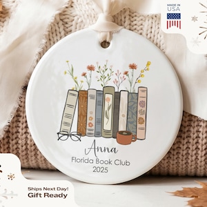 May include: A round white ceramic ornament featuring a colorful illustration of a row of books with wildflowers growing from the top, a pair of glasses, and a coffee mug. The text reads Anna, Florida Book Club, and 2025 in black lettering.