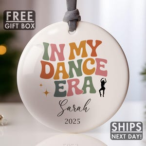 May include: White ceramic ornament with the text "IN MY DANCE ERA" in retro colors, a dancer silhouette, the name "Sarah", and the year 2025. The ornament has a gray ribbon for hanging. A "FREE GIFT BOX" and "SHIPS NEXT DAY!" graphic are also visible.