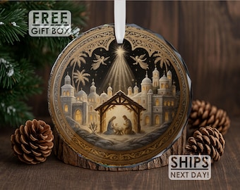 May include: Round Christmas ornament with a Nativity scene. The ornament features a detailed illustration of the Holy Family in a stable, with angels and a star. The ornament is trimmed in gold and has a white ribbon for hanging. The words "FREE GIFT BOX" are visible.