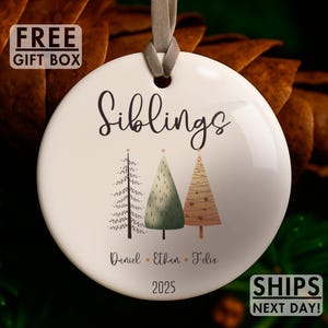 May include: A white ceramic Christmas ornament with the word "Siblings" in black script. It features three stylized Christmas trees in green and brown, with names and the year 2025 below. A gray ribbon is attached.