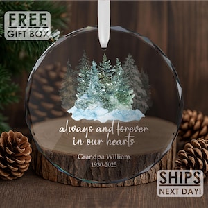 May include: A clear, round glass ornament with a winter forest scene and the text "always and forever in our hearts." The ornament rests on a wooden base and is surrounded by pine cones. The text "Grandpa William 1930-2025" is also visible.