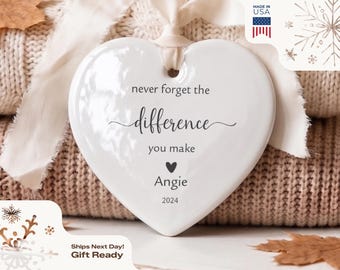 May include: A white heart-shaped ceramic ornament featuring the black printed text never forget the difference you make, a small heart icon, the name Angie, and the year 2024. A cream-colored ribbon is threaded through a hole at the top.