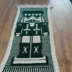 May include: A green and white crocheted table runner with a nutcracker design. The runner has a white fringe.