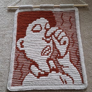 Puede incluir: Un tapiz de pared tejido a crochet con un diseño marrón y blanco. El diseño es de un fantasma con una cabeza grande y un cuerpo pequeño.