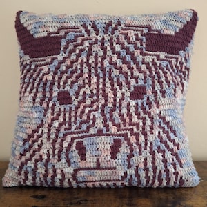 Op de afbeelding: Een vierkant gehaakt kussen met een Highland koeienontwerp. Het kussen heeft een textuur met een kleurenpalet van bordeauxrood, lichtblauw en roze. Het gezicht van de koe is prominent aanwezig, met hoorns, ogen en een snuit.