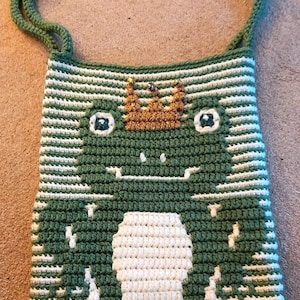 May include: A green and white striped crocheted bag featuring a frog design. The frog is wearing a gold crown with glitter. The bag has a long green strap.