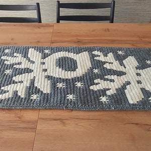 Op de afbeelding: Een geweven tafelkleed in grijs en wit met een sneeuwvlokkenpatroon. De tafelkleed ligt op een houten tafel.