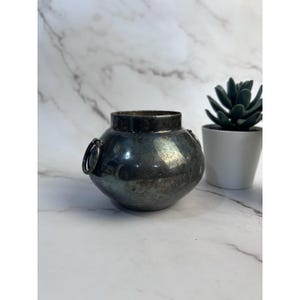 Op de afbeelding: Een kleine, ronde, zilverkleurige metalen vaas met twee ringhandvatten. De vaas heeft een donkere, verouderde uitstraling en staat op een wit marmeren oppervlak. Naast de vaas staat een kleine vetplant in pot.