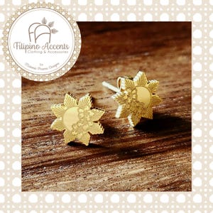 May include: Gold-colored stud earrings in a sunburst design with floral accents. The earrings are on a wooden surface. The earrings are from Filipino Accents Clothing & Accessories by Melanie Perone Designs.