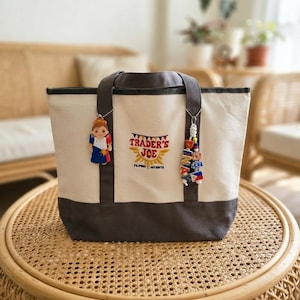 May include: A cream-colored canvas tote bag with dark gray trim and handles. The front features embroidered text reading TRADER'S JOE above FILIPINO ACCENTS. Two charms hang from the handles: one a figure in a traditional dress, the other a beaded keychain.