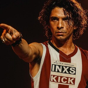 May include: A man wearing a red and white striped sleeveless jersey with the words "INXS KICK" printed on the front. He has dark curly hair and is pointing with his right hand. He is wearing a black bracelet.