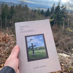Könnte beinhalten: Ein Buch mit dem Titel "KREUZE IM ERZGEBIRGE" wird im Freien gehalten. Das Cover zeigt ein Foto eines Kreuzes auf einem Feld unter blauem Himmel. Der Name des Autors, Kristin Richter, ist unten auf dem Cover abgedruckt.