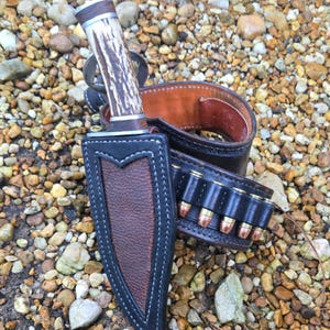 May include: A fixed-blade knife with a textured antler handle and a silver bolster, housed in a brown and black leather sheath with white stitching. Beside it lies a matching black leather belt featuring a series of brass-cased cartridges.
